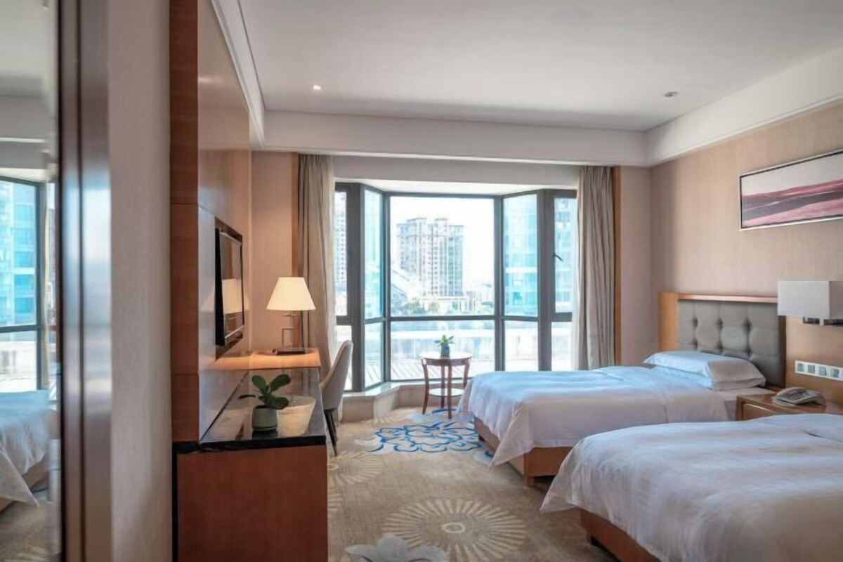 A hotel room with two beds, a desk with a lamp, a chair, and a view of the city through large windows.
