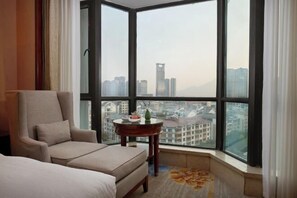 A hotel room with a large window offering a city view, a comfortable armchair, a small round table with a vase, and a patterned rug.