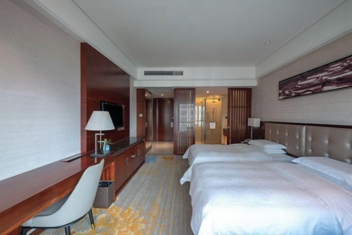 A hotel room with a desk, two beds, a TV, and a large painting on the wall.