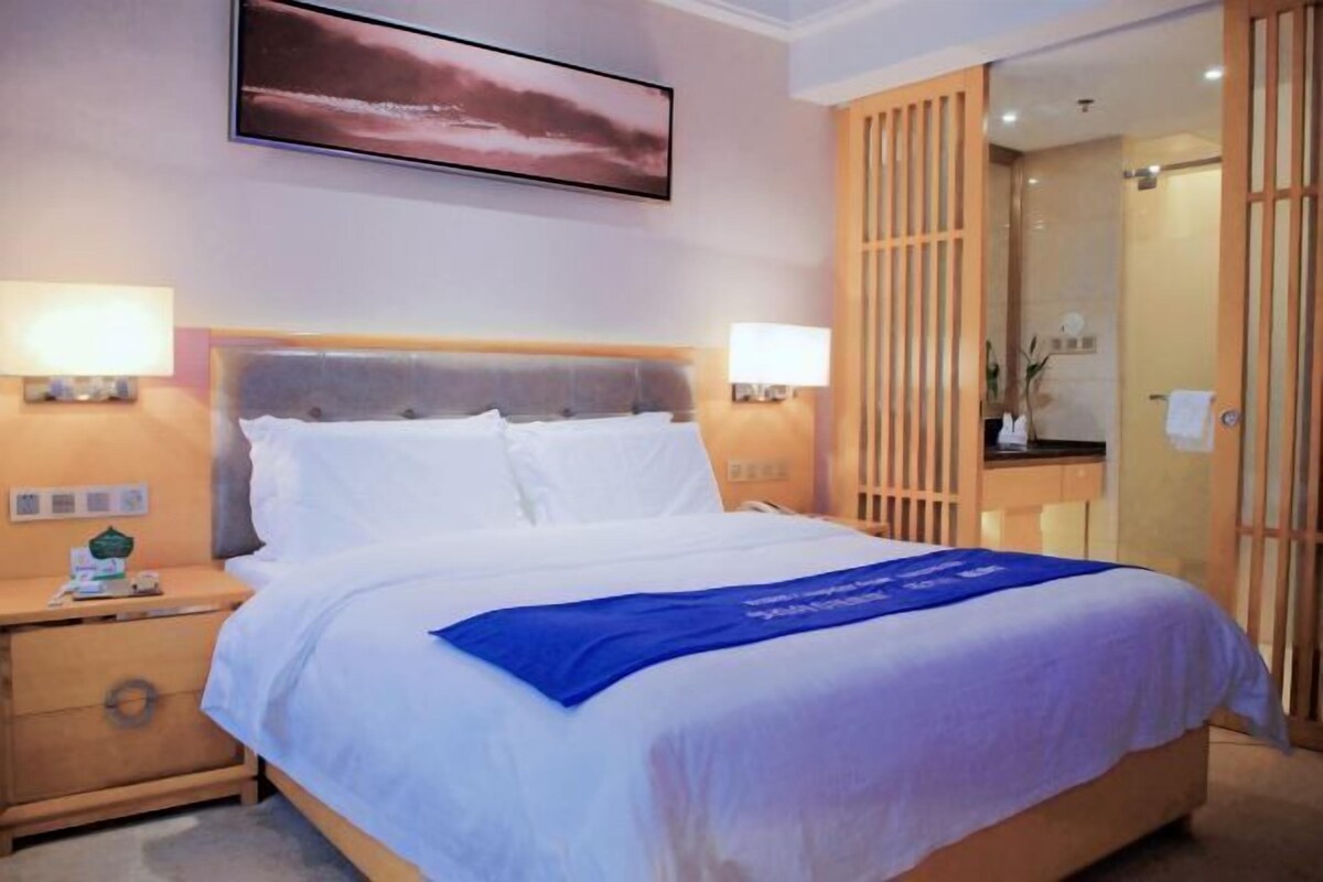 A hotel room with a bed, bedside lamps, a nightstand, a wooden wardrobe, and a painting on the wall.