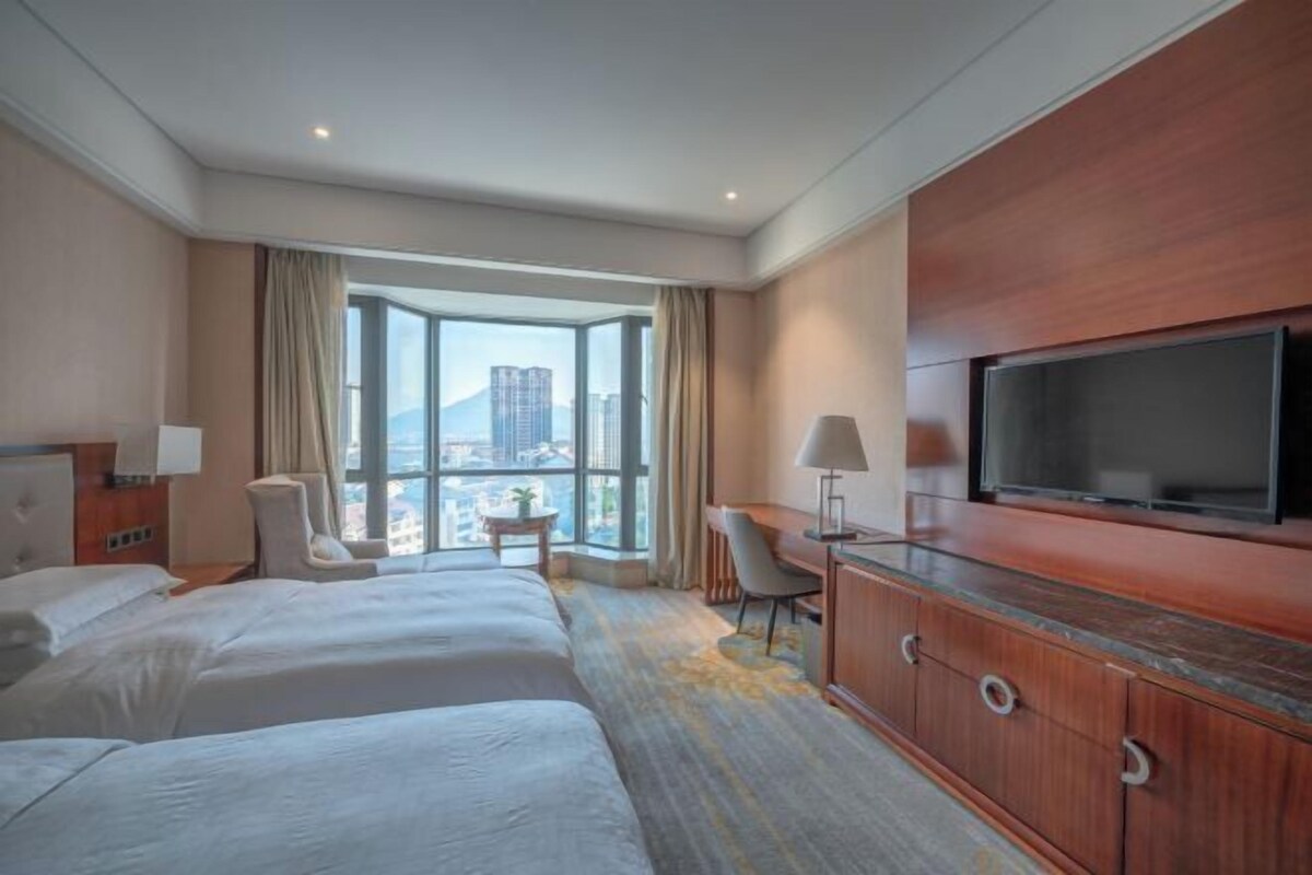 A hotel room with two beds, a desk, a television, and a large window offering a city view.