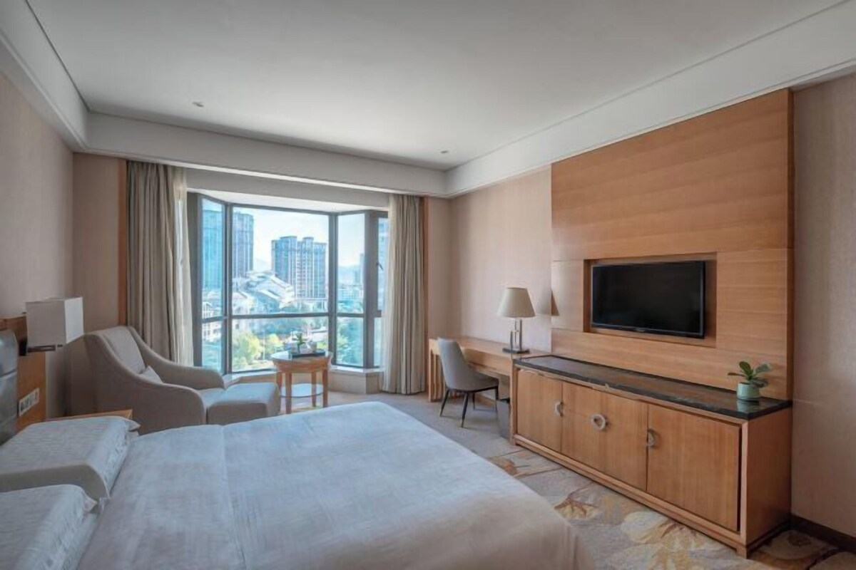 A hotel room with a large bed, a desk, a TV, and a view of the city through large windows.