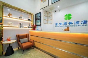 A reception area with a wooden reception desk, a brown chair, a small table with a red object, and shelves with decorative items. - Changtai Hezheng Business Hotel (Zhangzhou)