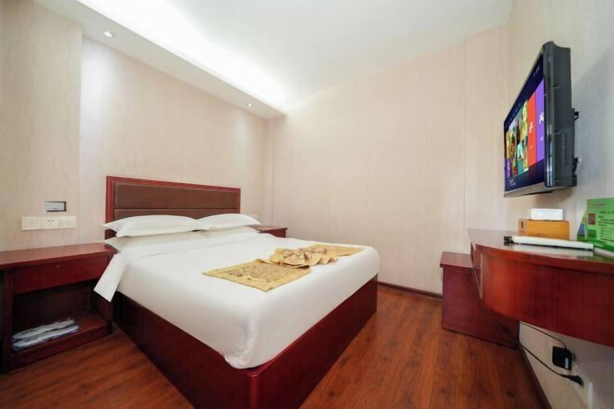A hotel room with a bed, wooden headboard, bedside tables, a desk with a chair, a flat-screen TV, and a small shelf.