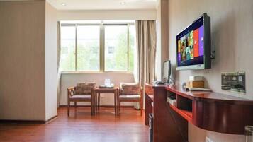 Una habitación de hotel con suelo de madera, un escritorio con computadora, un televisor montado en la pared y una ventana con cortinas.