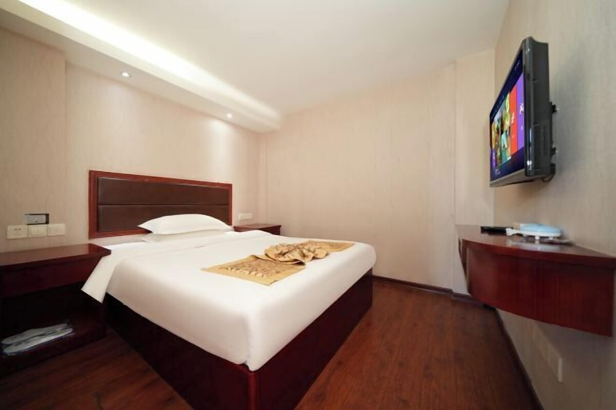 A hotel room with a wooden bed, a flat-screen TV mounted on the wall, a bedside table, and a wooden floor.