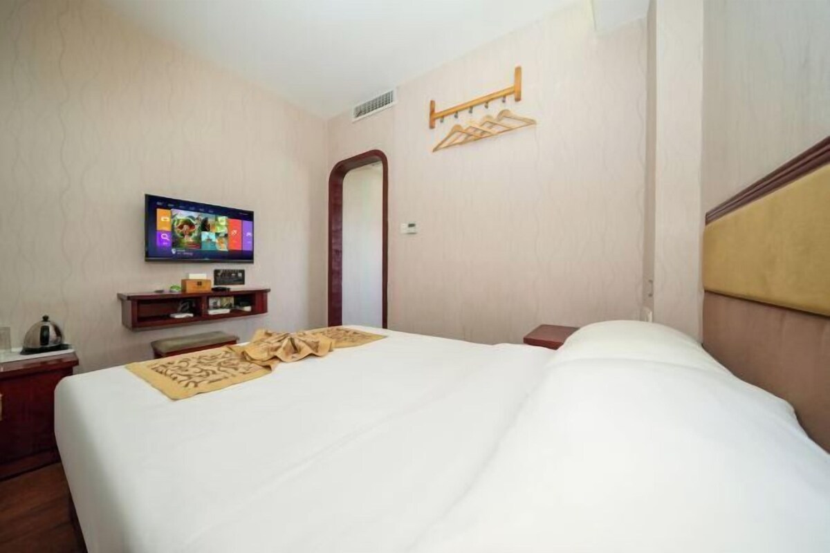A hotel room with a bed, a television on the wall, a wooden shelf with items, and a coat rack.