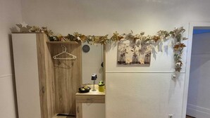 Quarto compacto com um guarda-roupa de madeira, uma pequena mesa de estudo com luminária e uma prateleira fixada na parede com potes e folhas decorativas.