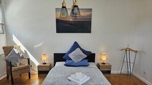 Quarto com cama, duas mesas de cabeceira com luminárias, uma cadeira, luminária pendente e um cabideiro.