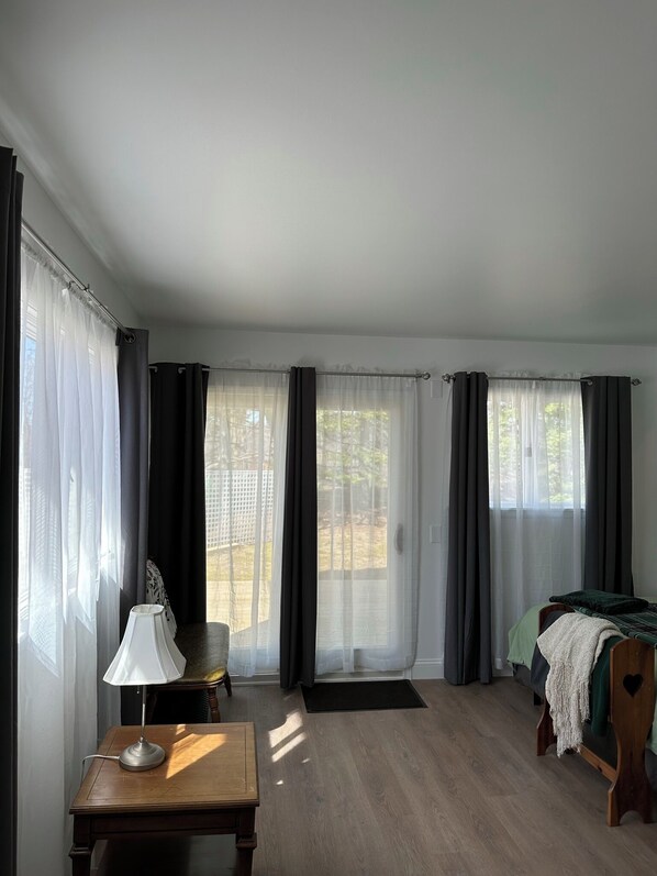 A room with a bed, a desk with a lamp, and large windows with curtains.