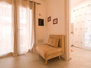 A small, beige, cushioned chair in a room with a tiled floor, a white curtain, and a door with a patterned curtain.