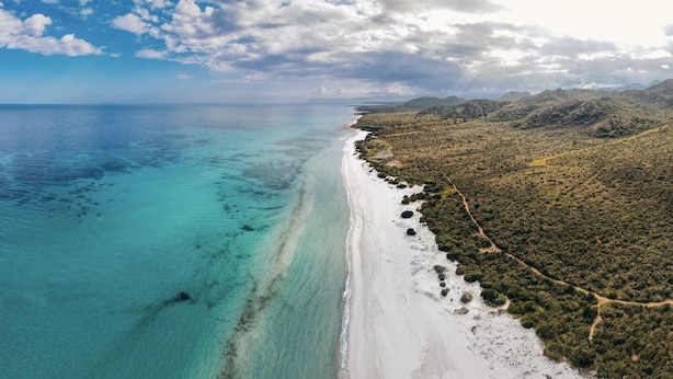 An aerial view of a beach with clear turquoise waters and a white sandy shore, surrounded by dense green vegetation and hills in the background.