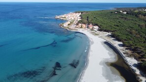 Vista aerea di una cittadina costiera con spiaggia, acque blu cristalline e una zona collinare con edifici.