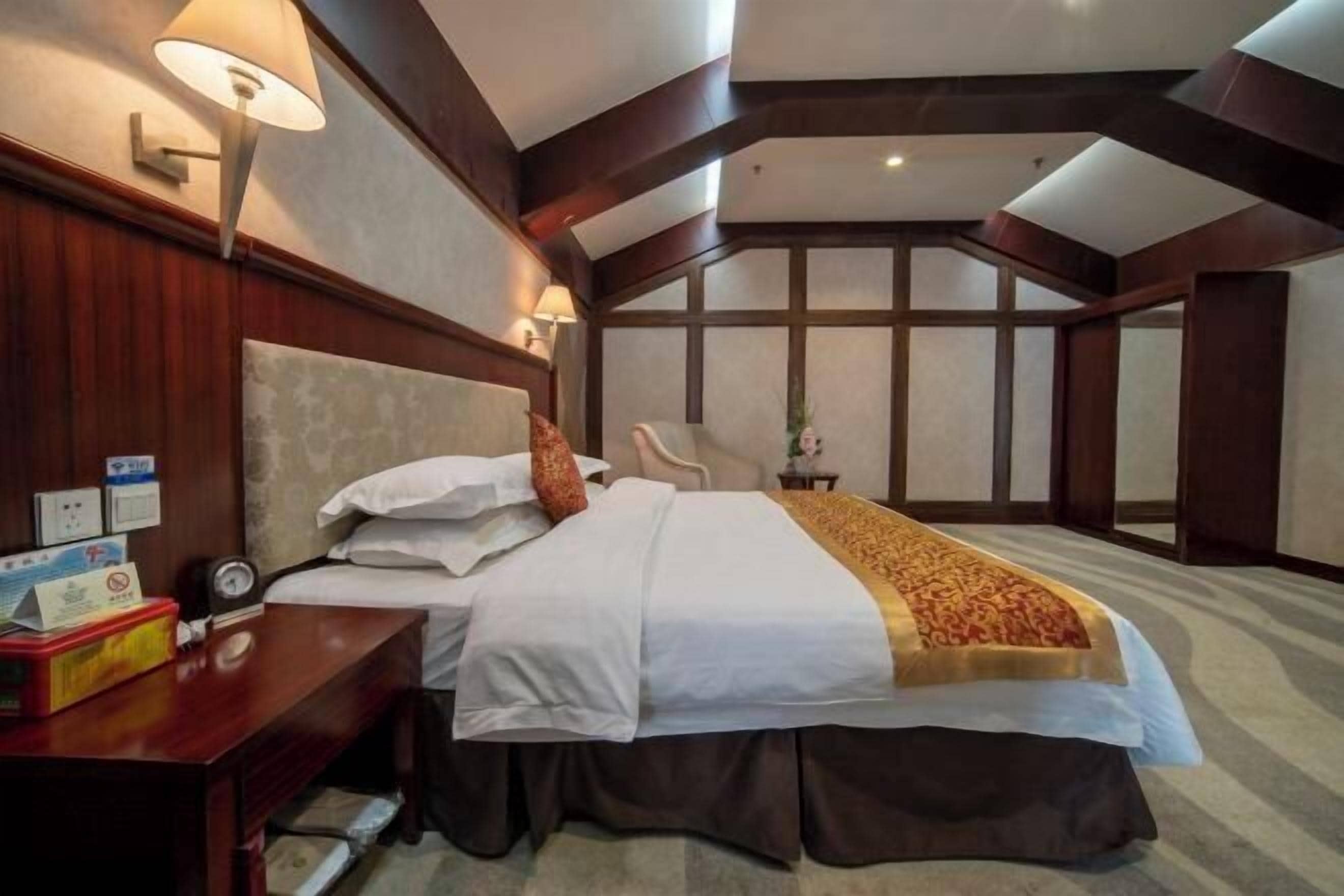 A hotel room with a large bed, a desk with a lamp, a chair, and a view of the sky through the window.