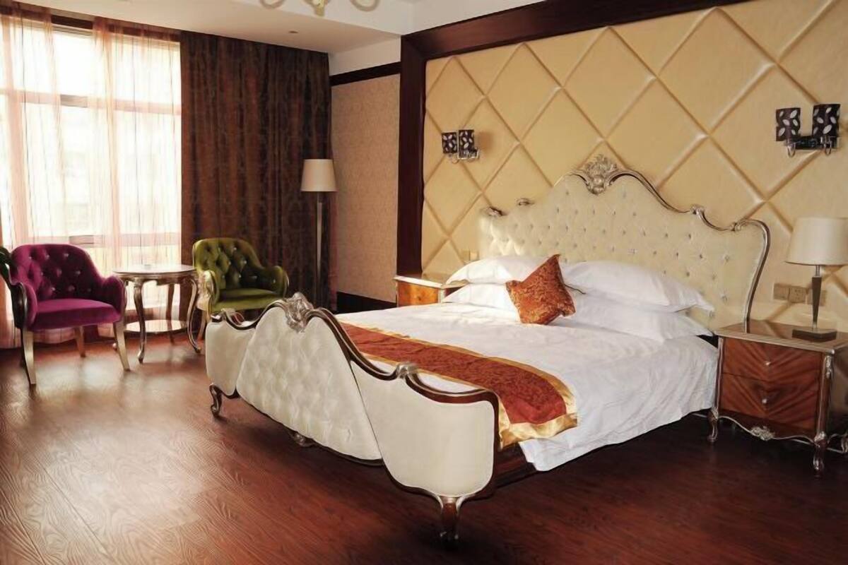 A hotel room with a large bed, a green armchair, a small round table, and a window with curtains.