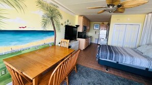 Een kamer met een houten eettafel, stoelen, een bed, een plafondventilator, een kitchenette en een muurschildering van een strand.