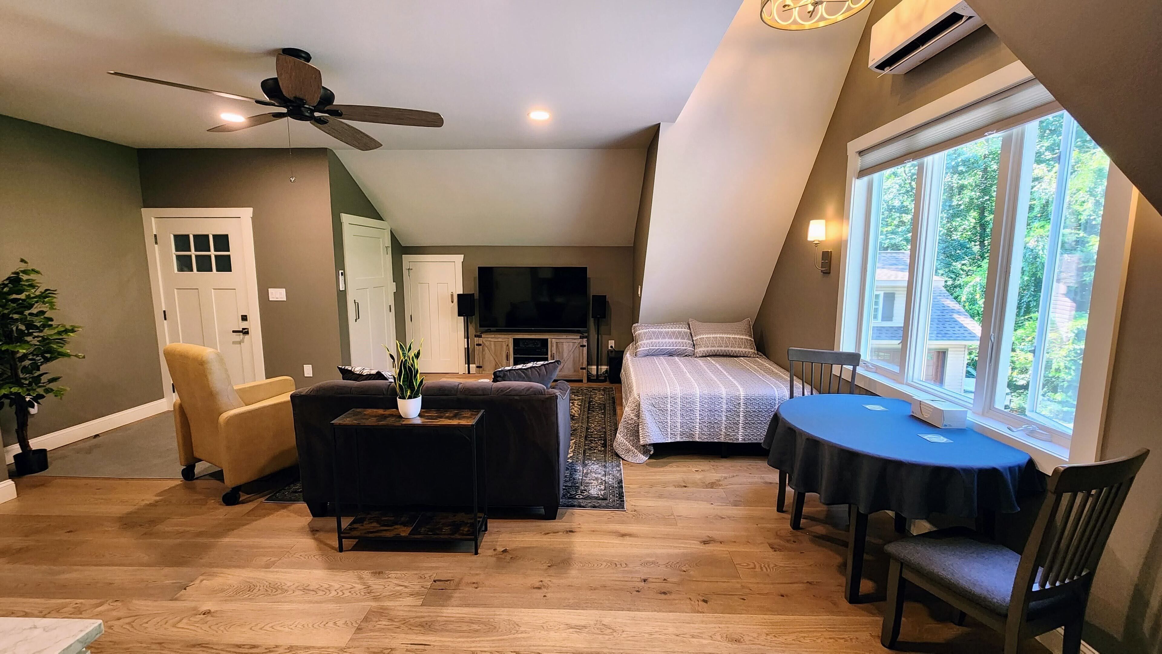A cozy loft bedroom with a ceiling fan, a large window, a bed, a TV, and a dining area with a table and chairs.