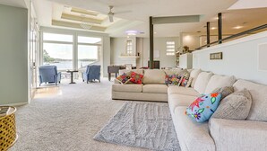 A spacious living room with a large beige sofa, a grey rug, and a view of the outdoors through large windows. - Exquisite Canal-Front Home: Access to Lake Conroe! (Montgomery)