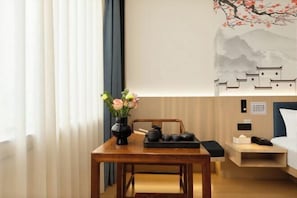 A hotel room with a wooden table, a vase with flowers, a bed, a nightstand, and a wall painting.