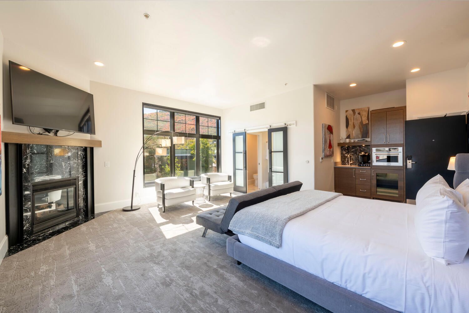 A modern bedroom with a large bed, a TV, a fireplace, and a view of the outdoors.