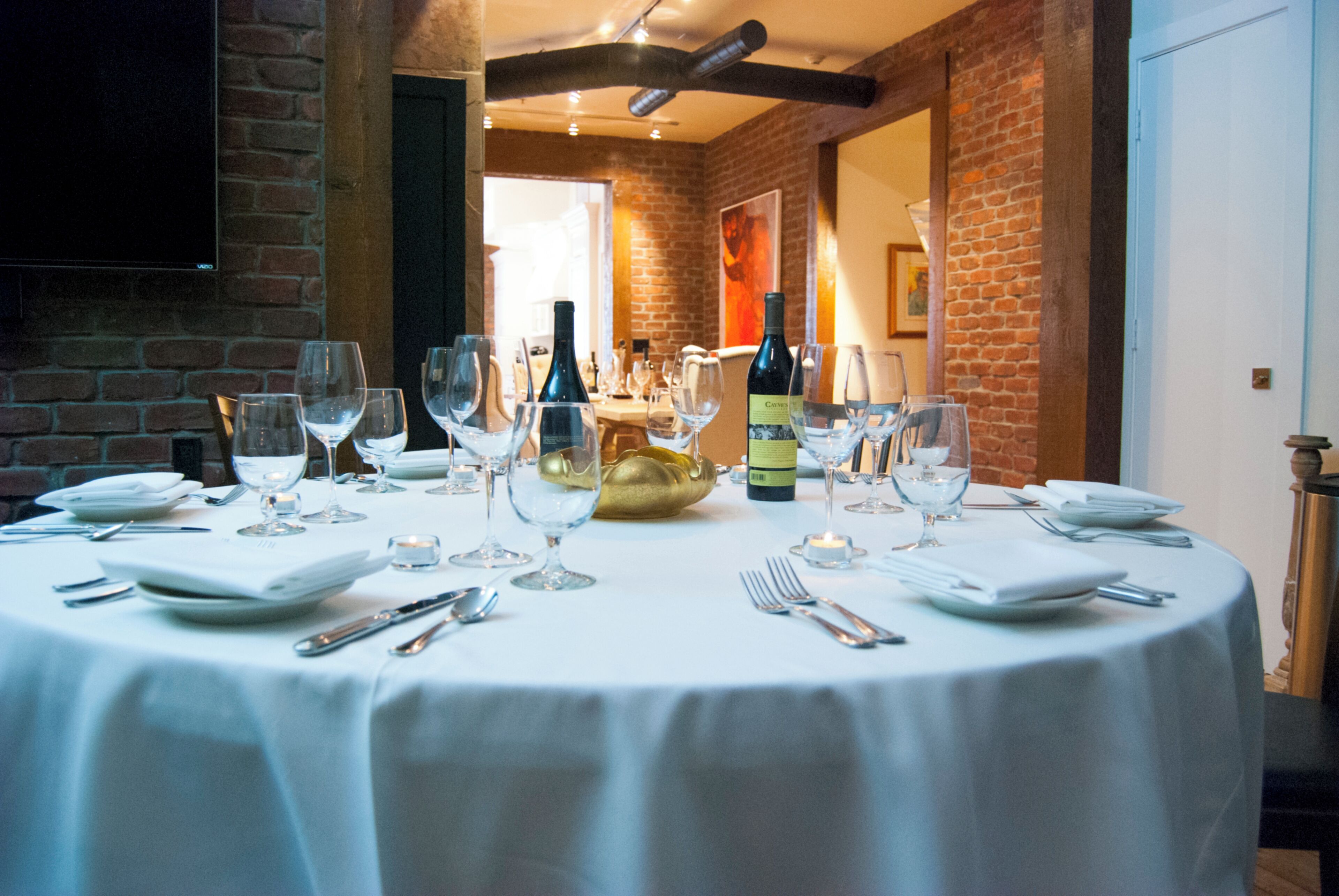 Une table de salle à manger ronde, recouverte d’une nappe blanche, avec des verres à vin, des assiettes, des couverts et une bouteille de vin.