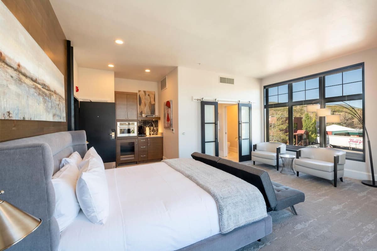 A modern hotel room with a large bed, a kitchenette, a seating area with a table and chairs, and a large window offering a view of the outside.