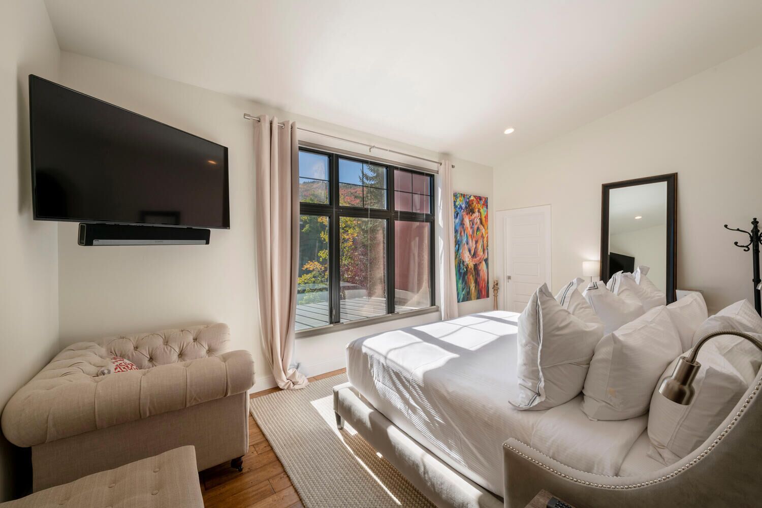 A modern bedroom with a large bed, a flat-screen TV, a sofa, and a window with a view of trees.