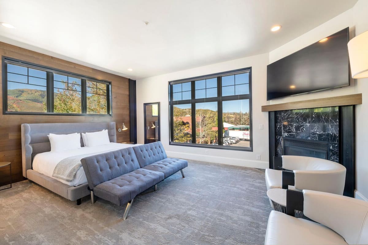 A modern bedroom with a large bed, a sofa, a TV, and large windows offering a view of the outdoors.