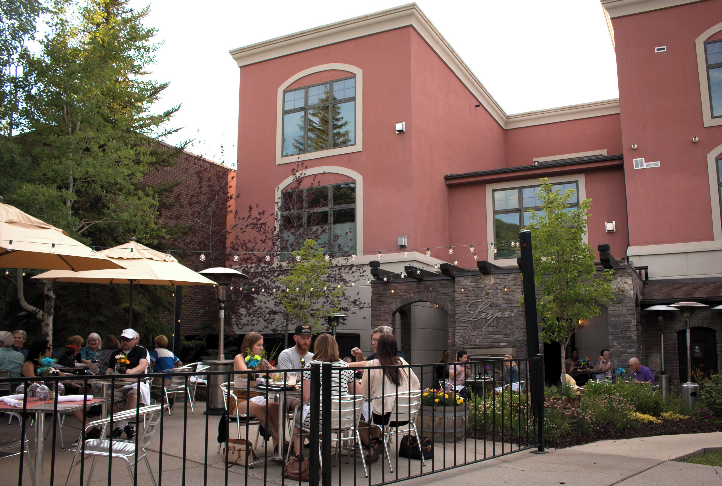 An outdoor patio area with tables and chairs, surrounded by a stone wall and a building with large windows and a brick facade.