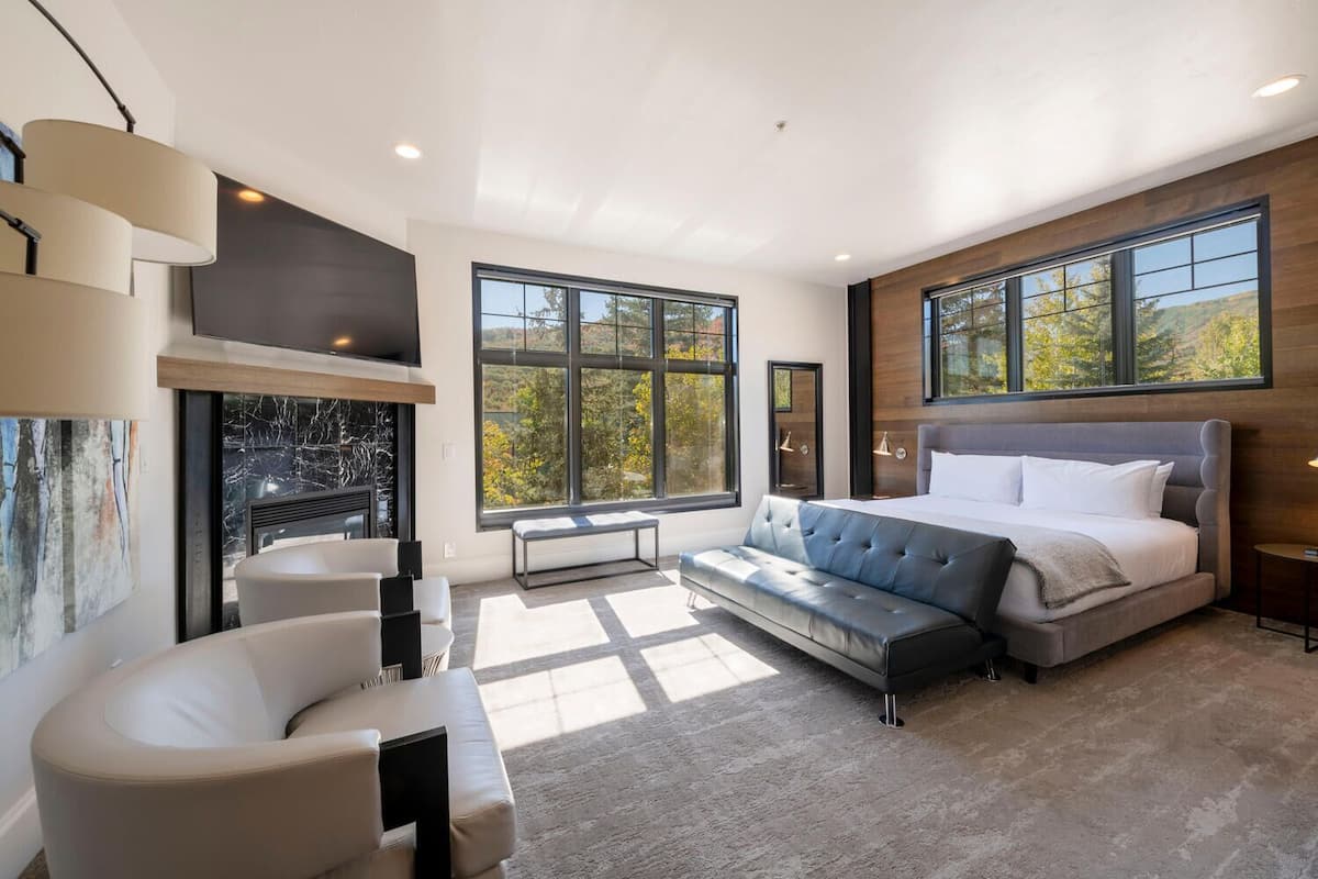 A modern bedroom with a large bed, a sofa, a fireplace, and large windows offering a view of trees.