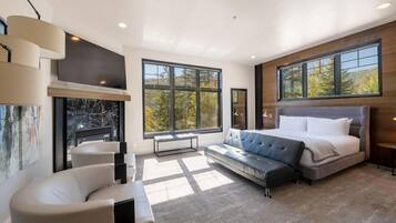 Een moderne slaapkamer met een groot bed, een bank, een open haard en grote ramen met uitzicht op bomen.