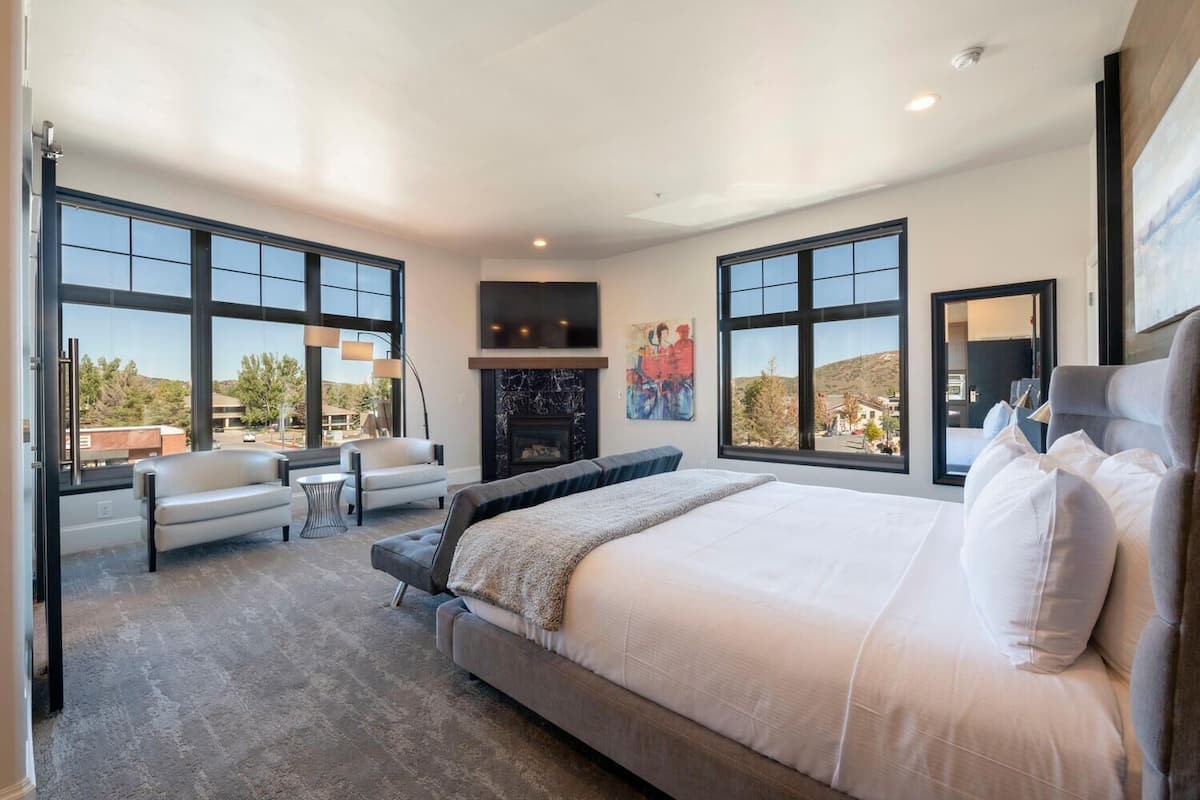 A modern bedroom with a large bed, two armchairs, a fireplace, and large windows offering a view of the outdoors.