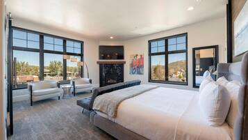 A modern bedroom with a large bed, two armchairs, a fireplace, and large windows offering a view of the outdoors.
