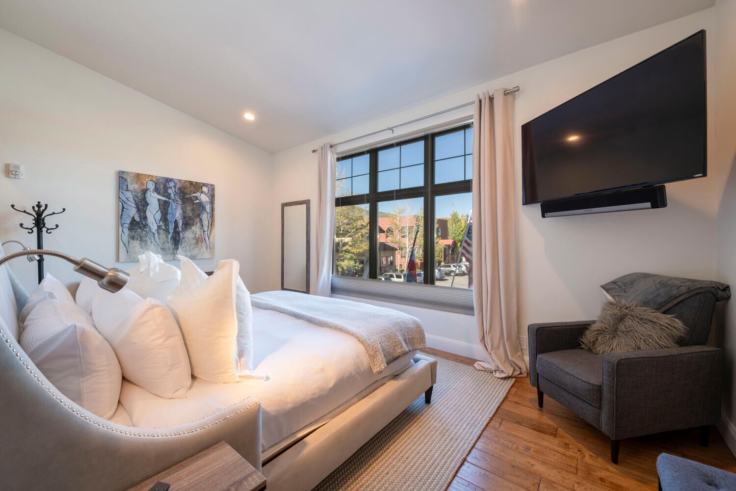 Un dormitorio moderno con una cama grande, un sillón gris, un televisor de pantalla plana y un ventanal con vistas al exterior.