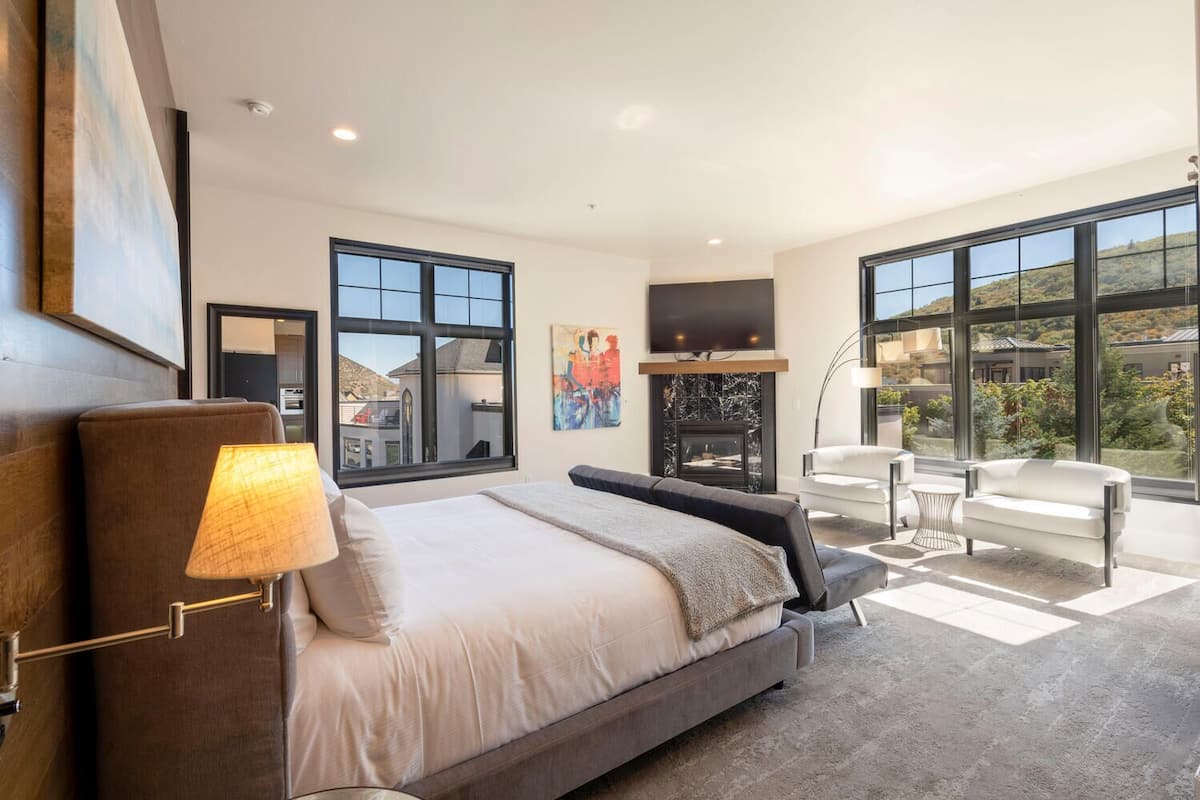A modern bedroom with a large bed, a fireplace, a TV, and large windows offering a view of the outdoors.