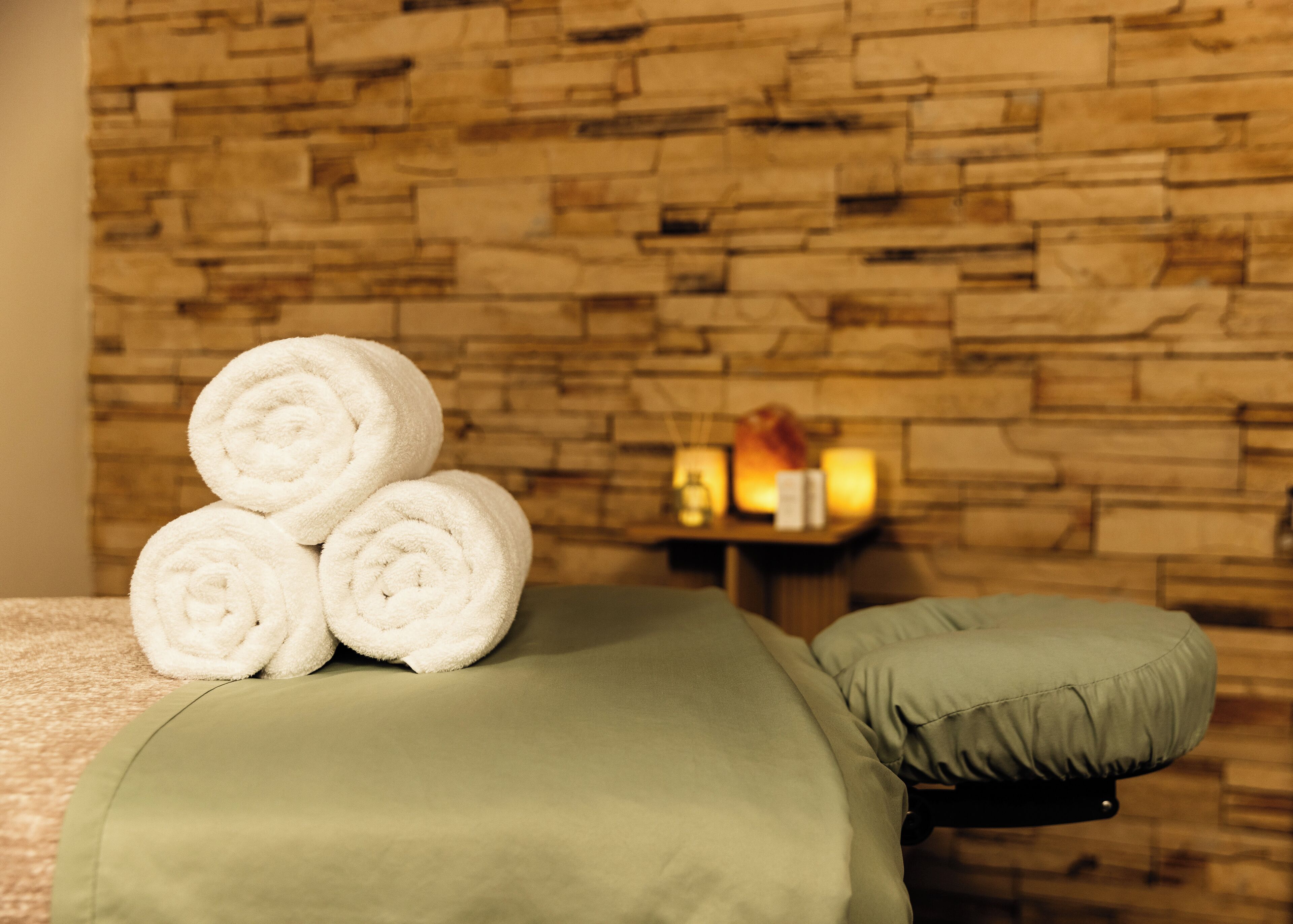 A spa room with a stone wall, a massage table with green cushions, and three rolled white towels.