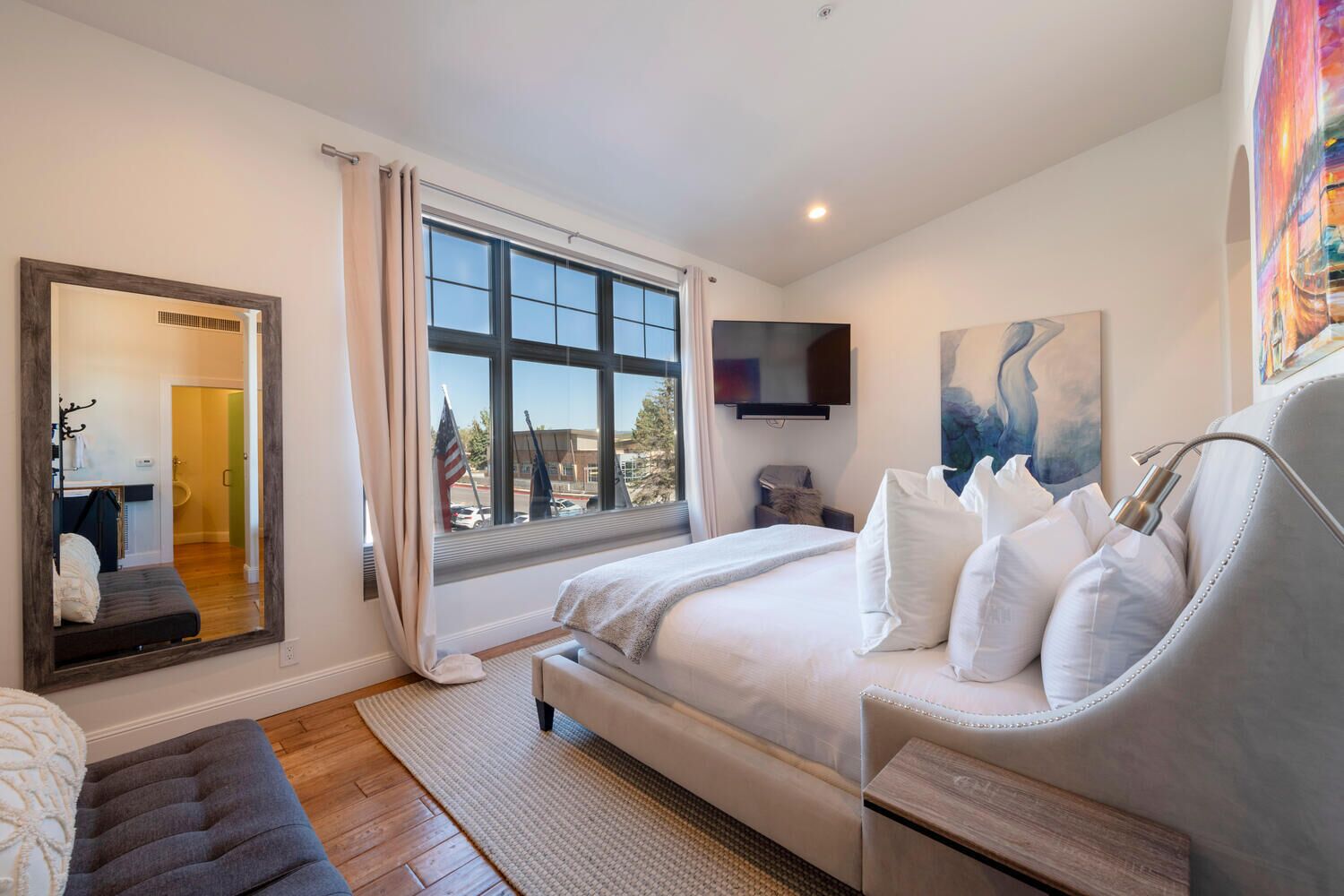 A modern bedroom with a large bed, a mirror, a TV, and a painting.