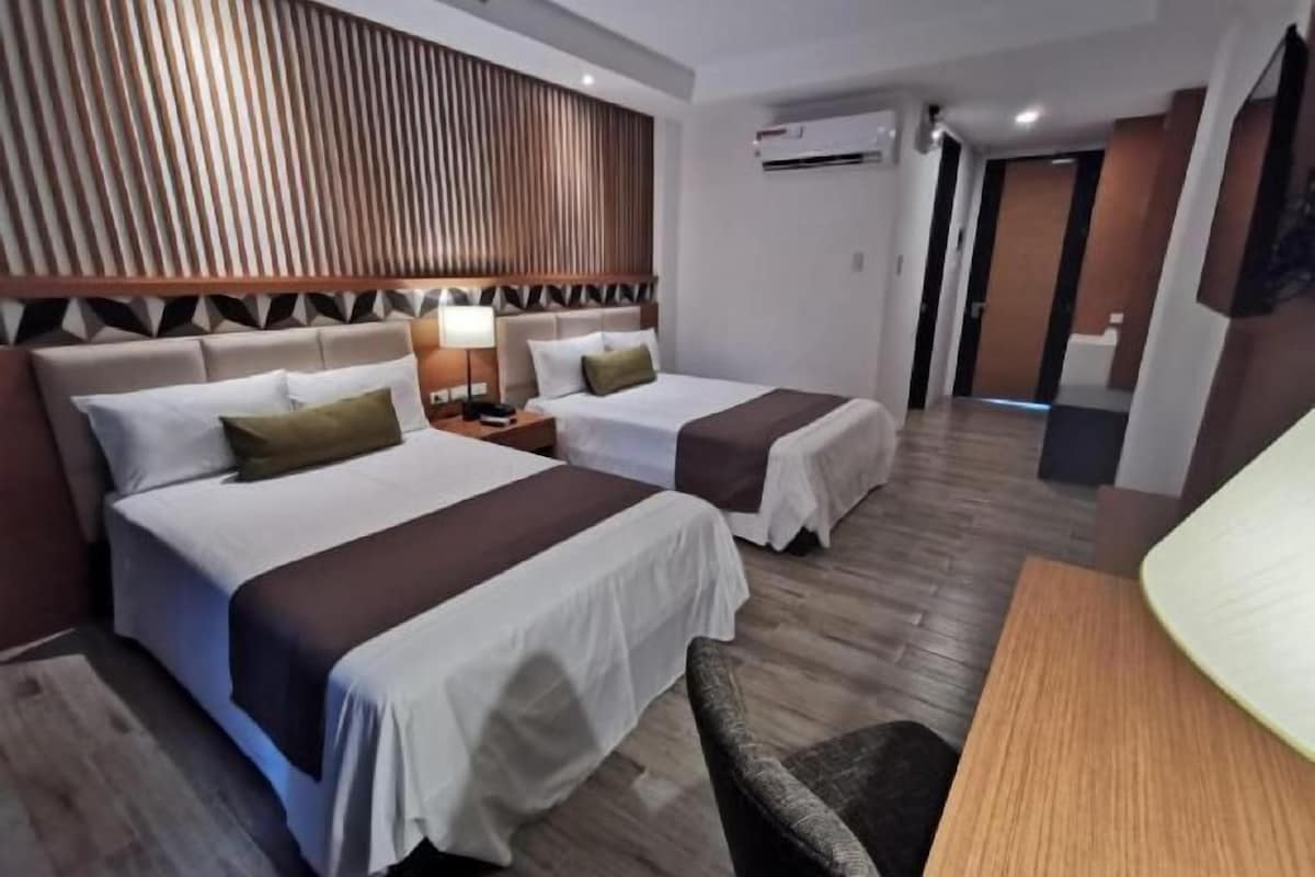 A hotel room with two beds, a wooden headboard, a bedside table with a lamp, and a small seating area.