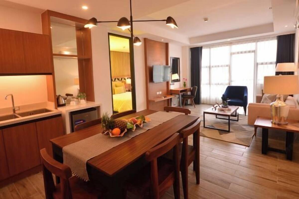 Ein modernes Hotelzimmer mit einem Holztisch, einer Küchenzeile, einer Sitzecke mit einer Couch und großen Fenstern.