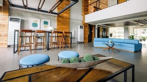 A modern lobby with a blue sofa, wooden chairs, a coffee table with greenery, and exposed beams. - Crown Hotel Palawan at Harbour Springs (Puerto Princesa)