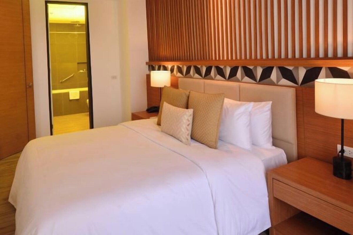 A hotel room with a large bed, wooden headboard, bedside lamps, and a visible bathroom.