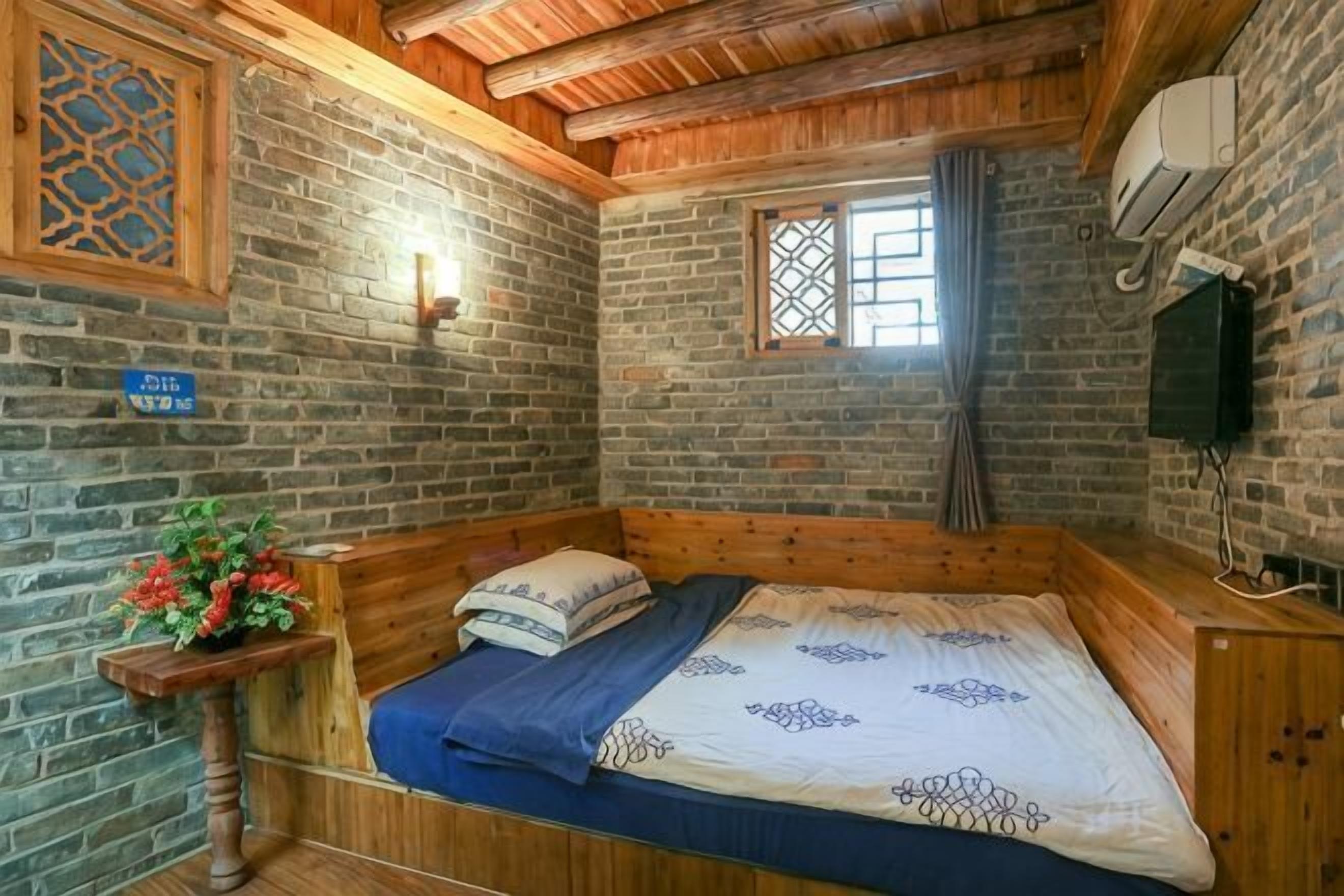 A wooden-framed room with a bed, a small table with a plant, a window with curtains, and a wall-mounted TV.