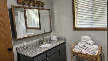 A bathroom with a granite countertop sink, dark cabinets, a large mirror, and a wicker chair with towels.