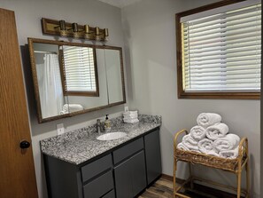 Un baño con lavamanos de encimera de granito, armarios oscuros, un espejo grande y una silla de mimbre con toallas.