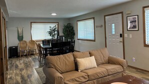 A living room with a beige sofa, a dining table with chairs, a ceiling fan, and a door with a picture frame.