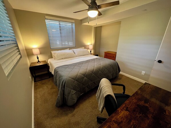 Un dormitorio con cama, mesita de noche, escritorio, silla, ventilador de techo y ventana con persianas.