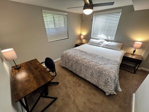 Un dormitorio con cama, dos mesitas de noche con lámparas, un escritorio con silla y dos ventanas con persianas.