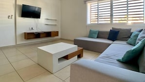 Una sala de estar moderna con un televisor de pantalla plana montado en la pared, una mesa de centro blanca, un sofá gris con varios cojines y una ventana con persianas.