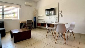 Un salon moderne comprenant un canapé, une table basse, une table à manger avec des chaises, un téléviseur fixé au mur et une fenêtre avec des stores.