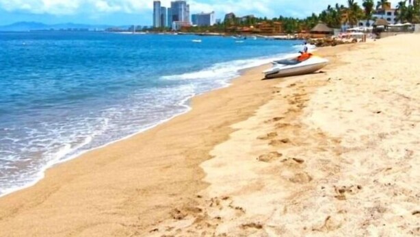 Une plage avec une eau d’un bleu limpide, un rivage sablonneux et une rangée de bâtiments en arrière-plan.